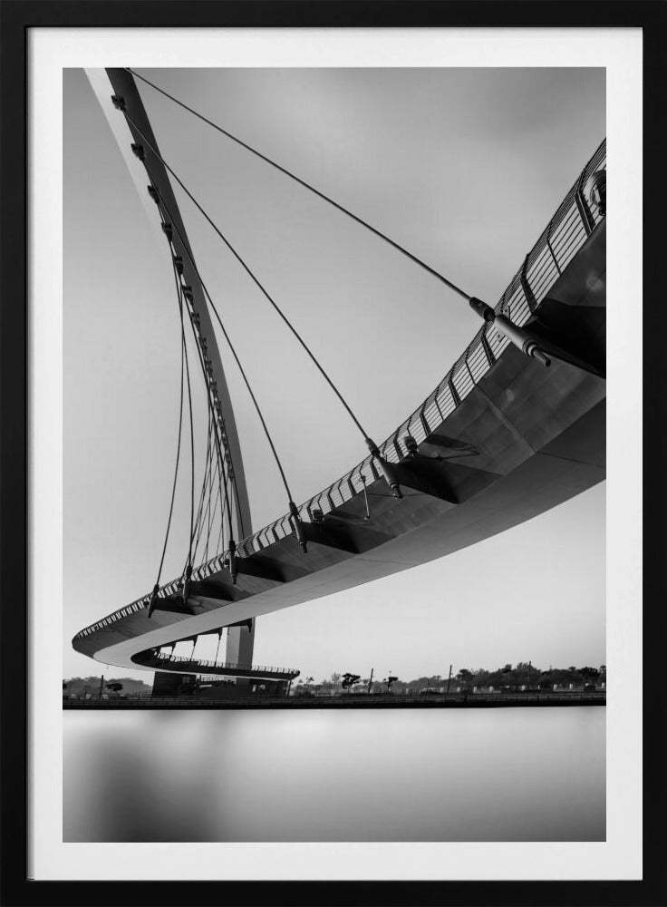 A dynamic low-angle, black and white photograph of a modern cable-stayed pedestrian bridge. The bridge curves elegantly over calm, reflective water, with its deck suspended by numerous cables attached to a tall, angled pylon, creating a striking graphic composition against a bright, clear sky. Decor