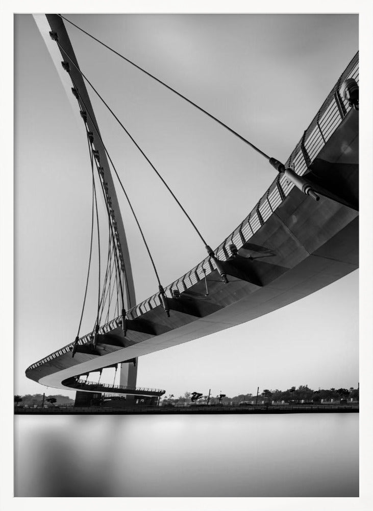 A dynamic low-angle, black and white photograph of a modern cable-stayed pedestrian bridge. The bridge curves elegantly over calm, reflective water, with its deck suspended by numerous cables attached to a tall, angled pylon, creating a striking graphic composition against a bright, clear sky. Decor