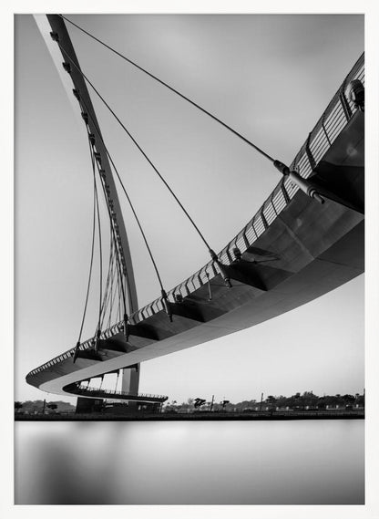 A dynamic low-angle, black and white photograph of a modern cable-stayed pedestrian bridge. The bridge curves elegantly over calm, reflective water, with its deck suspended by numerous cables attached to a tall, angled pylon, creating a striking graphic composition against a bright, clear sky. Decor