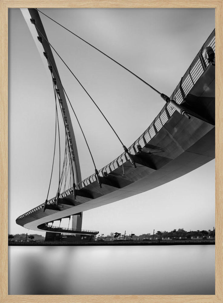 A dynamic low-angle, black and white photograph of a modern cable-stayed pedestrian bridge. The bridge curves elegantly over calm, reflective water, with its deck suspended by numerous cables attached to a tall, angled pylon, creating a striking graphic composition against a bright, clear sky. Decor