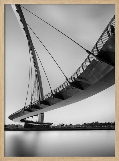 A dynamic low-angle, black and white photograph of a modern cable-stayed pedestrian bridge. The bridge curves elegantly over calm, reflective water, with its deck suspended by numerous cables attached to a tall, angled pylon, creating a striking graphic composition against a bright, clear sky. Decor