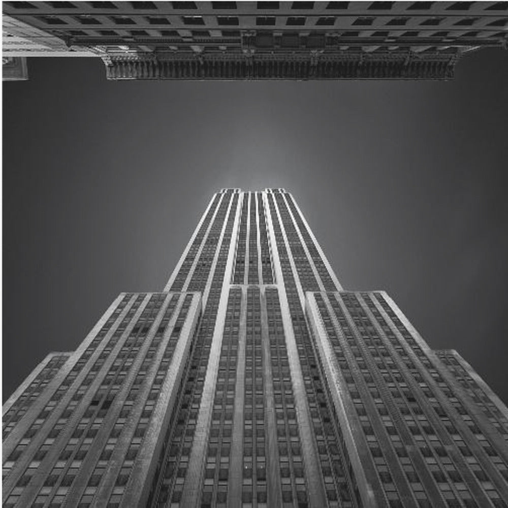 A dramatic, low-angle, black and white photograph of the Empire State Building, looking up from the street. The building's Art Deco lines stretch towards a dark grey sky, with another building visible at the very top of the frame, all enclosed in a light brown wood frame. Wall Art