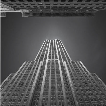 A dramatic, low-angle, black and white photograph of the Empire State Building, looking up from the street. The building's Art Deco lines stretch towards a dark grey sky, with another building visible at the very top of the frame, all enclosed in a light brown wood frame. Wall Art