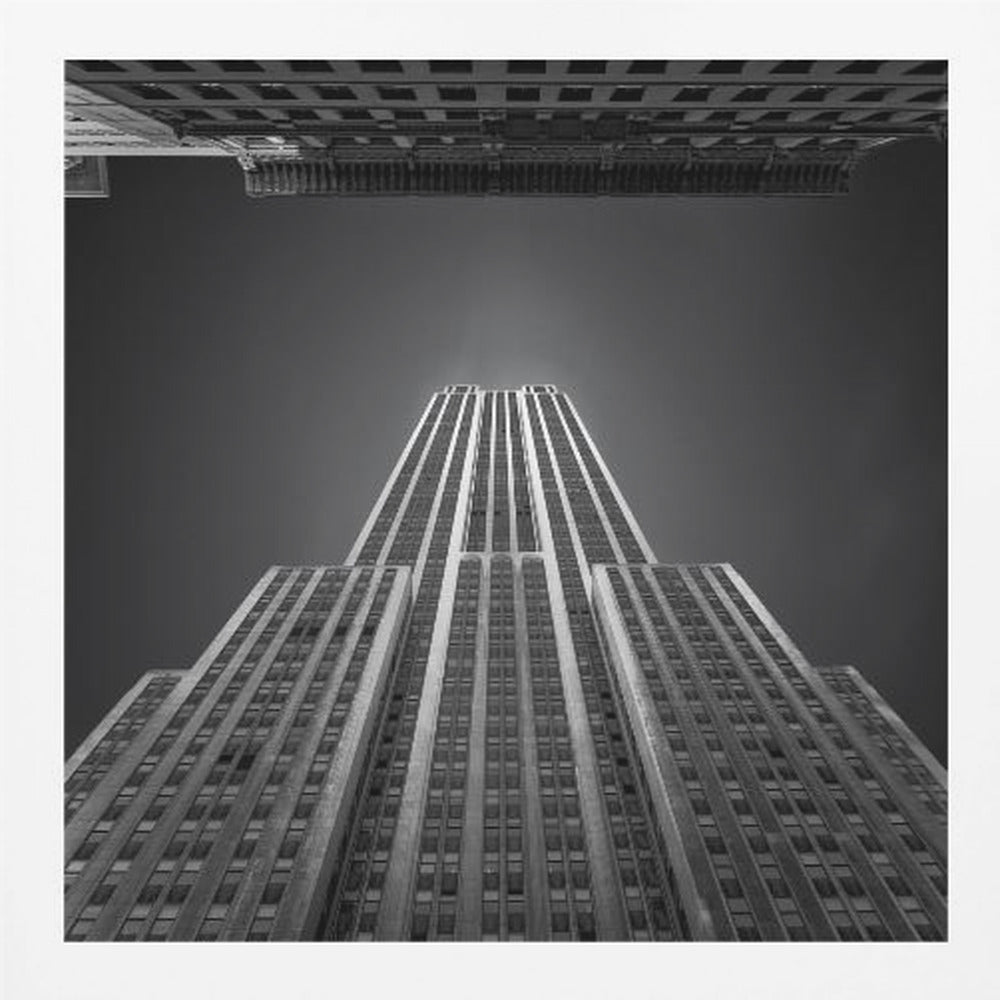 A dramatic, low-angle, black and white photograph of the Empire State Building, looking up from the street. The building's Art Deco lines stretch towards a dark grey sky, with another building visible at the very top of the frame, all enclosed in a light brown wood frame. Wall Art