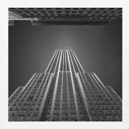 A dramatic, low-angle, black and white photograph of the Empire State Building, looking up from the street. The building's Art Deco lines stretch towards a dark grey sky, with another building visible at the very top of the frame, all enclosed in a light brown wood frame. Wall Art