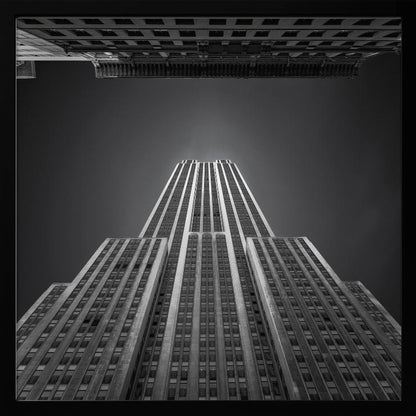 A dramatic, low-angle, black and white photograph of the Empire State Building, looking up from the street. The building's Art Deco lines stretch towards a dark grey sky, with another building visible at the very top of the frame, all enclosed in a light brown wood frame. Wall Art