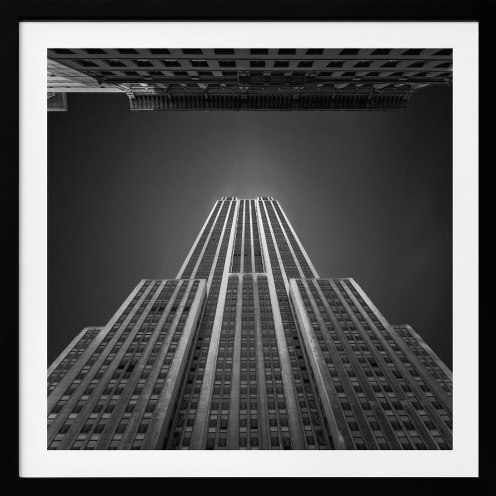A dramatic, low-angle, black and white photograph of the Empire State Building, looking up from the street. The building's Art Deco lines stretch towards a dark grey sky, with another building visible at the very top of the frame, all enclosed in a light brown wood frame. Wall Art