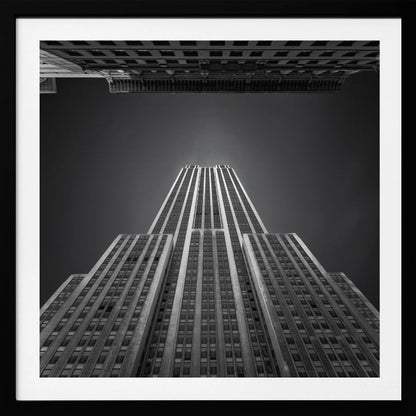 A dramatic, low-angle, black and white photograph of the Empire State Building, looking up from the street. The building's Art Deco lines stretch towards a dark grey sky, with another building visible at the very top of the frame, all enclosed in a light brown wood frame. Wall Art
