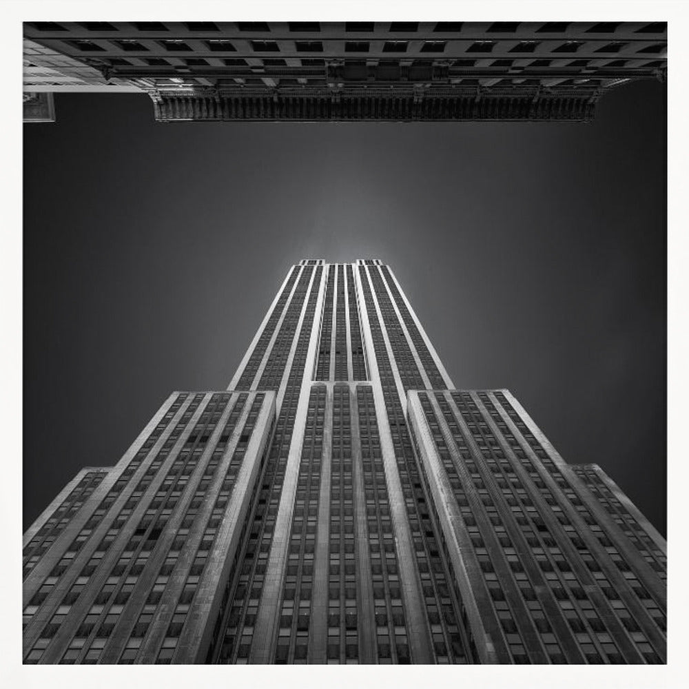 A dramatic, low-angle, black and white photograph of the Empire State Building, looking up from the street. The building's Art Deco lines stretch towards a dark grey sky, with another building visible at the very top of the frame, all enclosed in a light brown wood frame. Wall Art