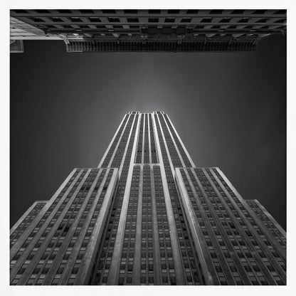 A dramatic, low-angle, black and white photograph of the Empire State Building, looking up from the street. The building's Art Deco lines stretch towards a dark grey sky, with another building visible at the very top of the frame, all enclosed in a light brown wood frame. Wall Art