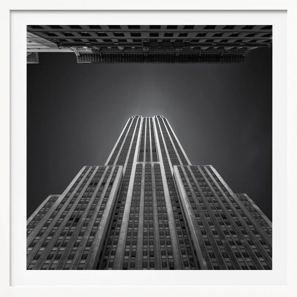 A dramatic, low-angle, black and white photograph of the Empire State Building, looking up from the street. The building's Art Deco lines stretch towards a dark grey sky, with another building visible at the very top of the frame, all enclosed in a light brown wood frame. Wall Art