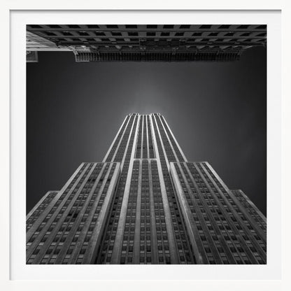 A dramatic, low-angle, black and white photograph of the Empire State Building, looking up from the street. The building's Art Deco lines stretch towards a dark grey sky, with another building visible at the very top of the frame, all enclosed in a light brown wood frame. Wall Art