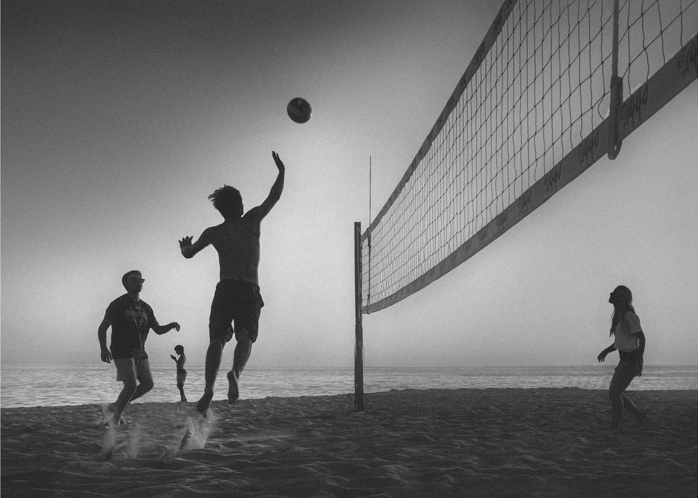 A black and white action shot of people playing volleyball on a beach, presented in a silver frame. The scene is silhouetted against a light sky, likely during sunrise or sunset. One player is captured mid-jump, arm raised to hit the ball over the net. Other players are visible on the sand, with the ocean in the background. Decor