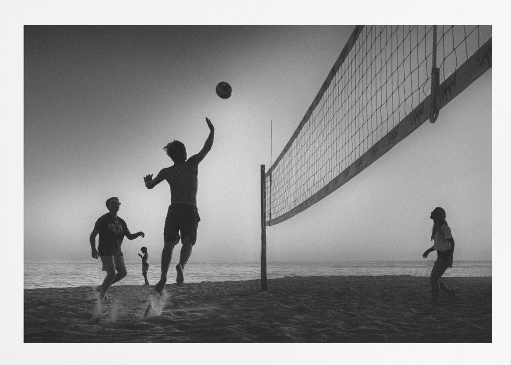 A black and white action shot of people playing volleyball on a beach, presented in a silver frame. The scene is silhouetted against a light sky, likely during sunrise or sunset. One player is captured mid-jump, arm raised to hit the ball over the net. Other players are visible on the sand, with the ocean in the background. Decor