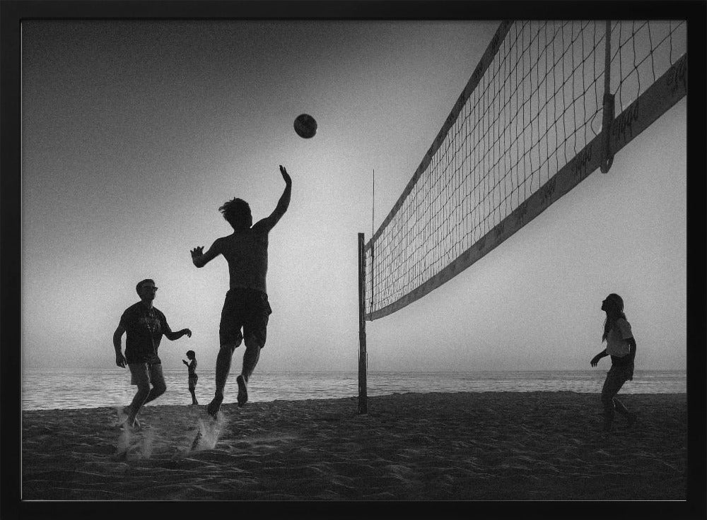 A black and white action shot of people playing volleyball on a beach, presented in a silver frame. The scene is silhouetted against a light sky, likely during sunrise or sunset. One player is captured mid-jump, arm raised to hit the ball over the net. Other players are visible on the sand, with the ocean in the background. Decor