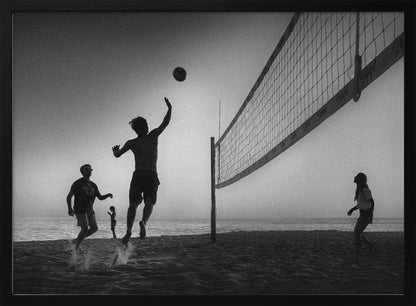 A black and white action shot of people playing volleyball on a beach, presented in a silver frame. The scene is silhouetted against a light sky, likely during sunrise or sunset. One player is captured mid-jump, arm raised to hit the ball over the net. Other players are visible on the sand, with the ocean in the background. Decor