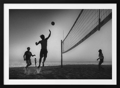 A black and white action shot of people playing volleyball on a beach, presented in a silver frame. The scene is silhouetted against a light sky, likely during sunrise or sunset. One player is captured mid-jump, arm raised to hit the ball over the net. Other players are visible on the sand, with the ocean in the background. Decor