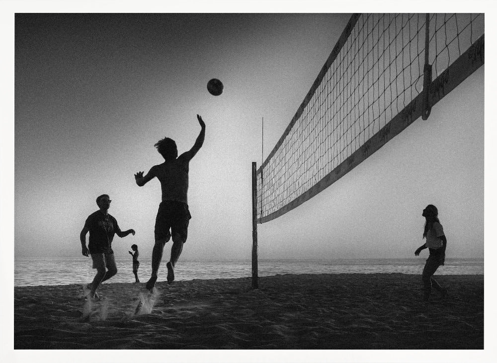 A black and white action shot of people playing volleyball on a beach, presented in a silver frame. The scene is silhouetted against a light sky, likely during sunrise or sunset. One player is captured mid-jump, arm raised to hit the ball over the net. Other players are visible on the sand, with the ocean in the background. Decor