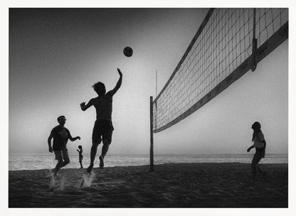 A black and white action shot of people playing volleyball on a beach, presented in a silver frame. The scene is silhouetted against a light sky, likely during sunrise or sunset. One player is captured mid-jump, arm raised to hit the ball over the net. Other players are visible on the sand, with the ocean in the background. Decor