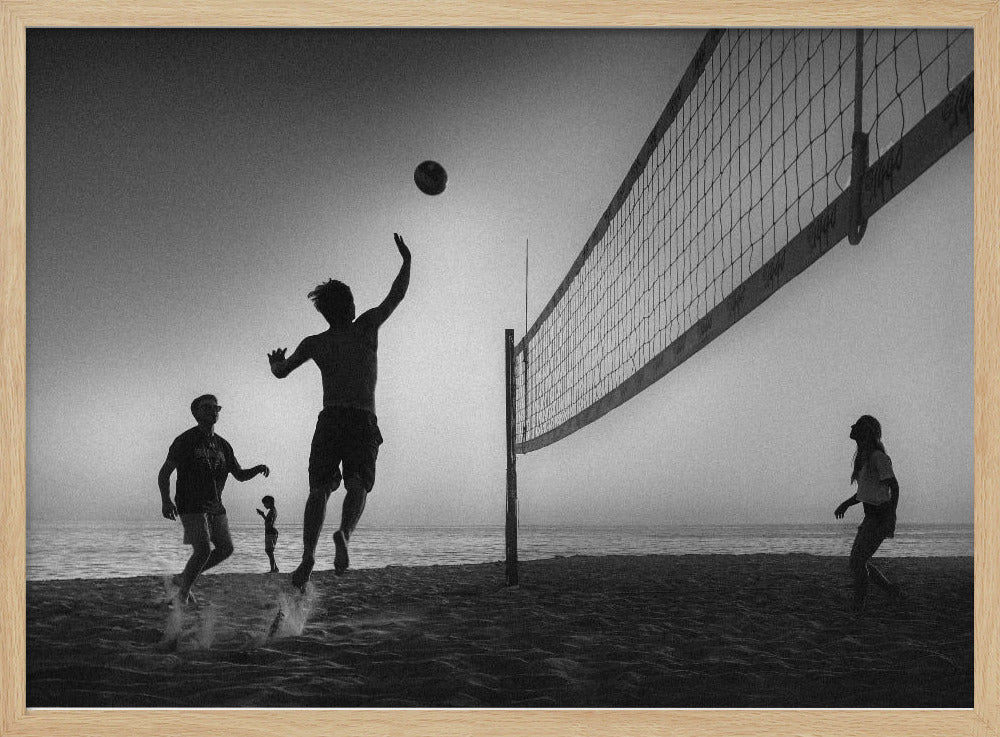 A black and white action shot of people playing volleyball on a beach, presented in a silver frame. The scene is silhouetted against a light sky, likely during sunrise or sunset. One player is captured mid-jump, arm raised to hit the ball over the net. Other players are visible on the sand, with the ocean in the background. Decor