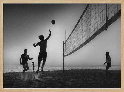 A black and white action shot of people playing volleyball on a beach, presented in a silver frame. The scene is silhouetted against a light sky, likely during sunrise or sunset. One player is captured mid-jump, arm raised to hit the ball over the net. Other players are visible on the sand, with the ocean in the background. Decor