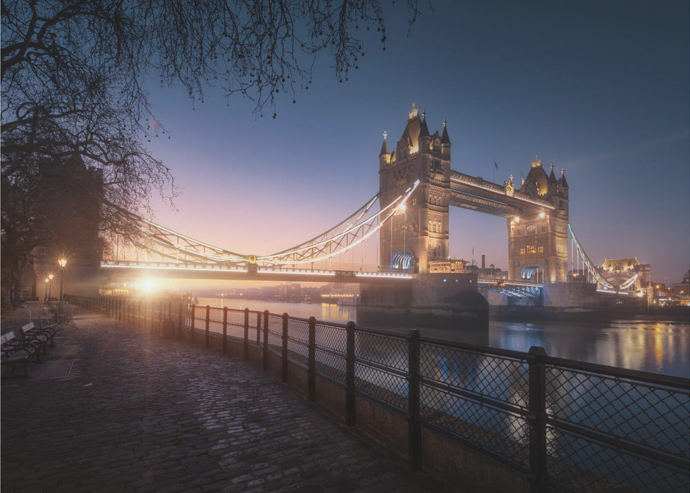 A scenic photograph of London's Tower Bridge at sunrise, its lights glowing warmly against a deep blue and orange sky. In the foreground, a cobblestone path follows the River Thames, leading the eye towards the iconic bridge and the rising sun. Poster