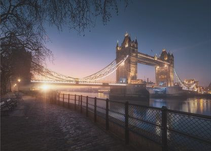 A scenic photograph of London's Tower Bridge at sunrise, its lights glowing warmly against a deep blue and orange sky. In the foreground, a cobblestone path follows the River Thames, leading the eye towards the iconic bridge and the rising sun. Poster