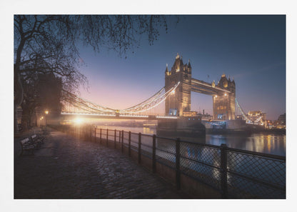 A scenic photograph of London's Tower Bridge at sunrise, its lights glowing warmly against a deep blue and orange sky. In the foreground, a cobblestone path follows the River Thames, leading the eye towards the iconic bridge and the rising sun. Poster