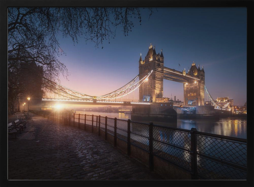 A scenic photograph of London's Tower Bridge at sunrise, its lights glowing warmly against a deep blue and orange sky. In the foreground, a cobblestone path follows the River Thames, leading the eye towards the iconic bridge and the rising sun. Poster