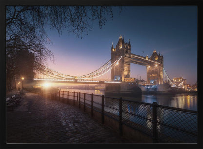 A scenic photograph of London's Tower Bridge at sunrise, its lights glowing warmly against a deep blue and orange sky. In the foreground, a cobblestone path follows the River Thames, leading the eye towards the iconic bridge and the rising sun. Poster
