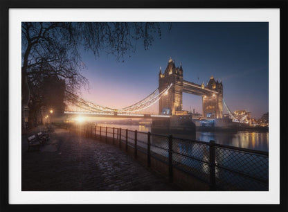 A scenic photograph of London's Tower Bridge at sunrise, its lights glowing warmly against a deep blue and orange sky. In the foreground, a cobblestone path follows the River Thames, leading the eye towards the iconic bridge and the rising sun. Poster