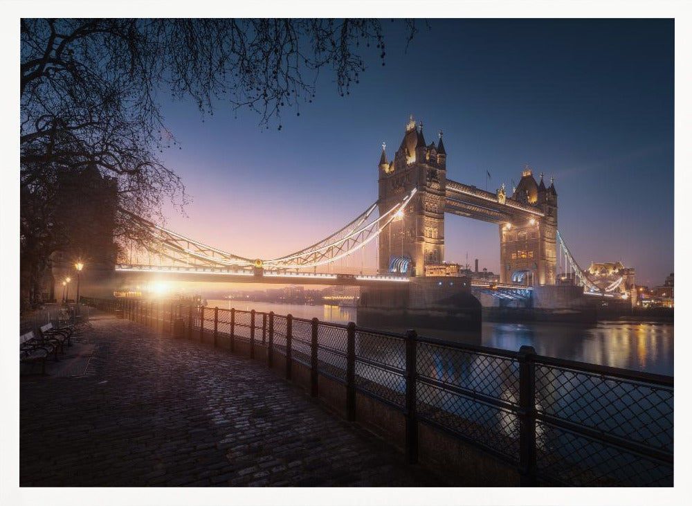 A scenic photograph of London's Tower Bridge at sunrise, its lights glowing warmly against a deep blue and orange sky. In the foreground, a cobblestone path follows the River Thames, leading the eye towards the iconic bridge and the rising sun. Poster