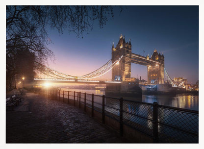 A scenic photograph of London's Tower Bridge at sunrise, its lights glowing warmly against a deep blue and orange sky. In the foreground, a cobblestone path follows the River Thames, leading the eye towards the iconic bridge and the rising sun. Poster