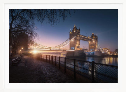 A scenic photograph of London's Tower Bridge at sunrise, its lights glowing warmly against a deep blue and orange sky. In the foreground, a cobblestone path follows the River Thames, leading the eye towards the iconic bridge and the rising sun. Poster