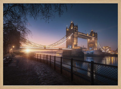 A scenic photograph of London's Tower Bridge at sunrise, its lights glowing warmly against a deep blue and orange sky. In the foreground, a cobblestone path follows the River Thames, leading the eye towards the iconic bridge and the rising sun. Poster