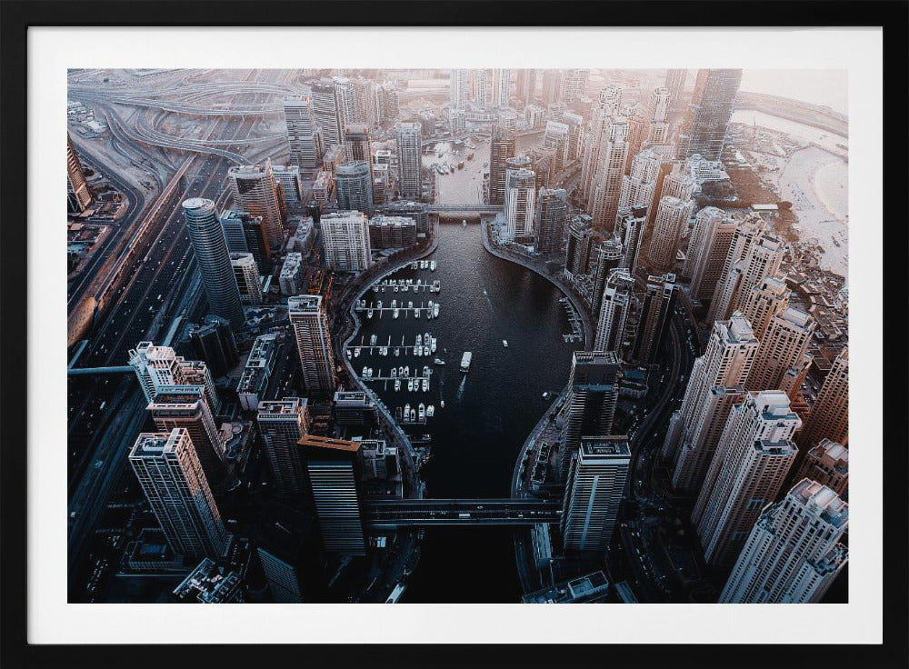 A high-angle aerial view of a dense, modern cityscape with a prominent dark-water marina shaped like a lightbulb. Numerous skyscrapers surround the marina, where many white boats are docked. To the left, a complex network of highways and overpasses weaves through the city. The photograph has a cool, desaturated color palette and is enclosed in a silver frame. Artwork