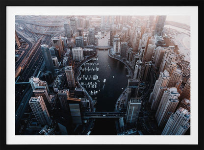 A high-angle aerial view of a dense, modern cityscape with a prominent dark-water marina shaped like a lightbulb. Numerous skyscrapers surround the marina, where many white boats are docked. To the left, a complex network of highways and overpasses weaves through the city. The photograph has a cool, desaturated color palette and is enclosed in a silver frame. Artwork