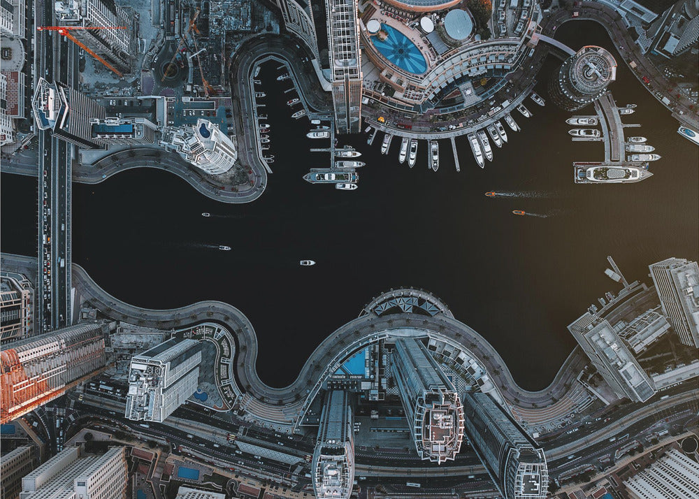A dramatic top-down aerial photograph of a sprawling modern city at night. A dark waterway curves through the center, surrounded by illuminated skyscrapers, busy roads, and a marina packed with boats, all within a silver picture frame. Artwork