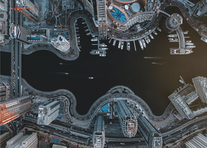 A dramatic top-down aerial photograph of a sprawling modern city at night. A dark waterway curves through the center, surrounded by illuminated skyscrapers, busy roads, and a marina packed with boats, all within a silver picture frame. Artwork