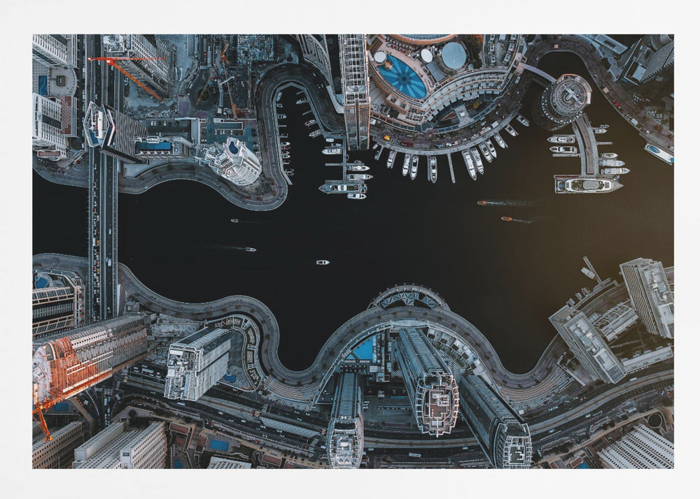 A dramatic top-down aerial photograph of a sprawling modern city at night. A dark waterway curves through the center, surrounded by illuminated skyscrapers, busy roads, and a marina packed with boats, all within a silver picture frame. Artwork