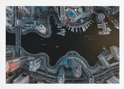 A dramatic top-down aerial photograph of a sprawling modern city at night. A dark waterway curves through the center, surrounded by illuminated skyscrapers, busy roads, and a marina packed with boats, all within a silver picture frame. Artwork