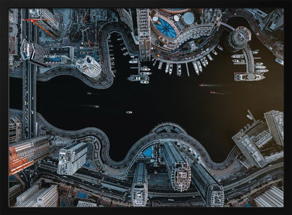 A dramatic top-down aerial photograph of a sprawling modern city at night. A dark waterway curves through the center, surrounded by illuminated skyscrapers, busy roads, and a marina packed with boats, all within a silver picture frame. Artwork