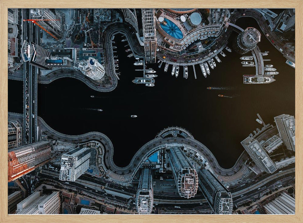 A dramatic top-down aerial photograph of a sprawling modern city at night. A dark waterway curves through the center, surrounded by illuminated skyscrapers, busy roads, and a marina packed with boats, all within a silver picture frame. Artwork