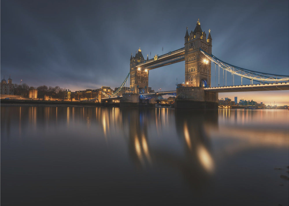 A long-exposure photograph of London's Tower Bridge at dusk, brilliantly illuminated with golden lights that cast shimmering reflections on the calm River Thames. The sky is a deep, dark blue, and the city skyline glows in the background, all enclosed within a silver frame. Print