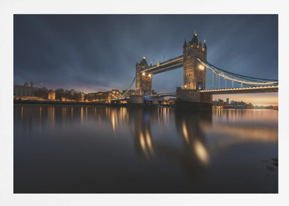 A long-exposure photograph of London's Tower Bridge at dusk, brilliantly illuminated with golden lights that cast shimmering reflections on the calm River Thames. The sky is a deep, dark blue, and the city skyline glows in the background, all enclosed within a silver frame. Print