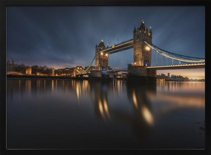 A long-exposure photograph of London's Tower Bridge at dusk, brilliantly illuminated with golden lights that cast shimmering reflections on the calm River Thames. The sky is a deep, dark blue, and the city skyline glows in the background, all enclosed within a silver frame. Print