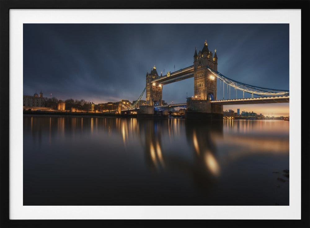 A long-exposure photograph of London's Tower Bridge at dusk, brilliantly illuminated with golden lights that cast shimmering reflections on the calm River Thames. The sky is a deep, dark blue, and the city skyline glows in the background, all enclosed within a silver frame. Print