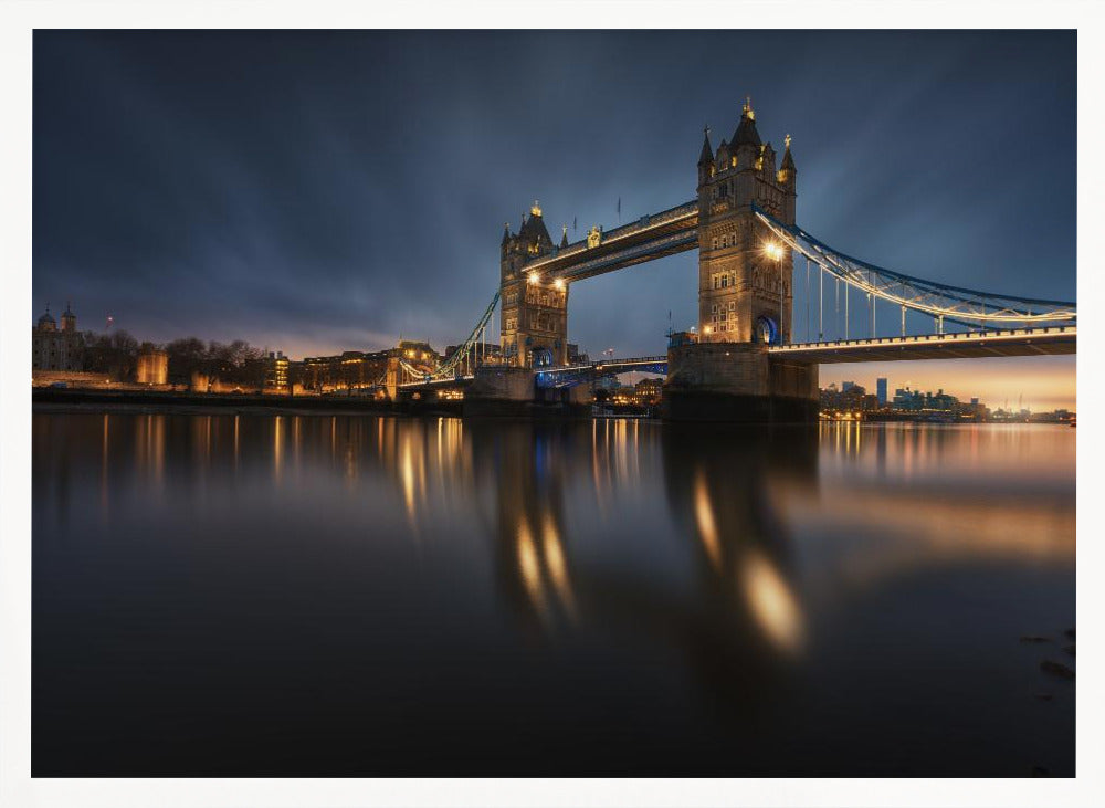 A long-exposure photograph of London's Tower Bridge at dusk, brilliantly illuminated with golden lights that cast shimmering reflections on the calm River Thames. The sky is a deep, dark blue, and the city skyline glows in the background, all enclosed within a silver frame. Print
