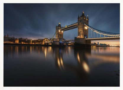 A long-exposure photograph of London's Tower Bridge at dusk, brilliantly illuminated with golden lights that cast shimmering reflections on the calm River Thames. The sky is a deep, dark blue, and the city skyline glows in the background, all enclosed within a silver frame. Print