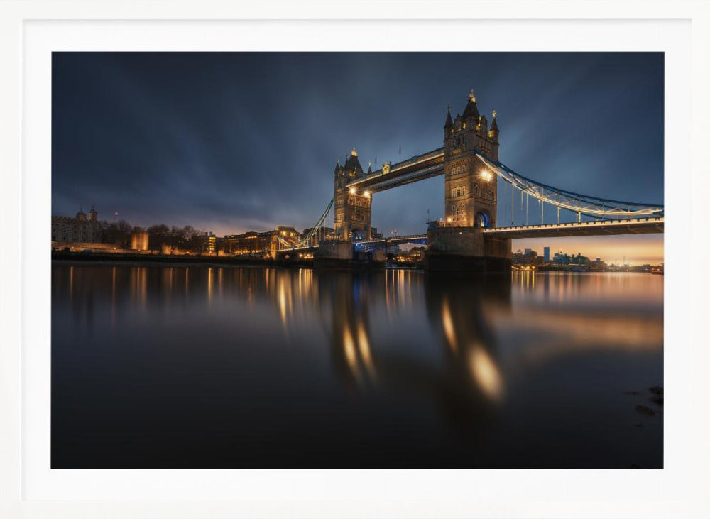 A long-exposure photograph of London's Tower Bridge at dusk, brilliantly illuminated with golden lights that cast shimmering reflections on the calm River Thames. The sky is a deep, dark blue, and the city skyline glows in the background, all enclosed within a silver frame. Print
